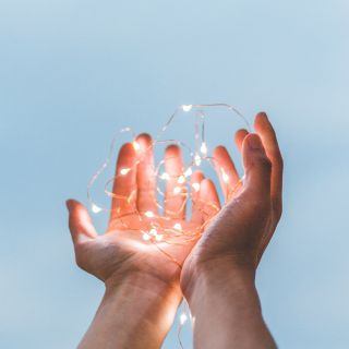 Mani che tengono fili di luci LED accese su sfondo cielo azzurro, simbolo di speranza e positività
