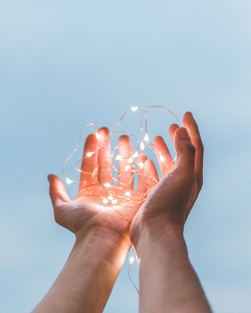 Mani che tengono fili di luci LED accese su sfondo cielo azzurro, simbolo di speranza e positività