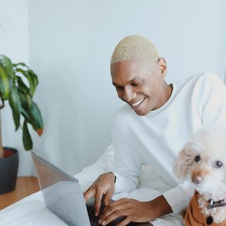 Uomo sorride lavorando al laptop su letto smart home, smartphone e cane accanto, ambiente moderno e luminoso