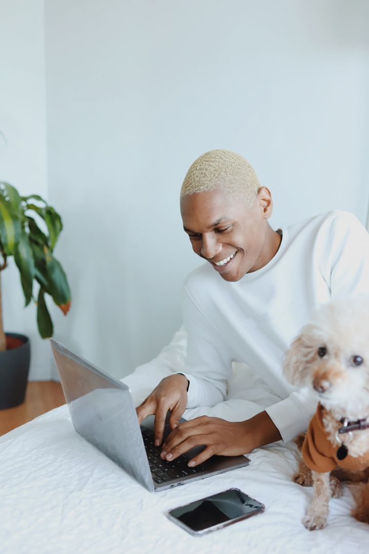 Uomo sorride lavorando al laptop su letto smart home, smartphone e cane accanto, ambiente moderno e luminoso