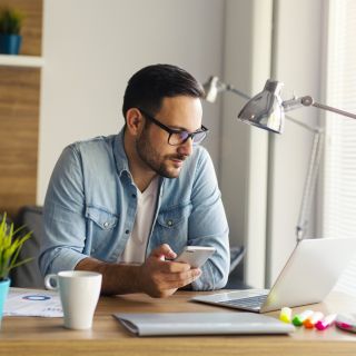 Uomo al computer che confronta offerte luce e gas per risparmiare sulla bolletta, smartphone e documenti su scrivania