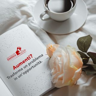 Tazza di caffè accanto a libro con scritta "Aumenti? Trasforma un problema in un’opportunità" e rosa bianca su lenzuolo