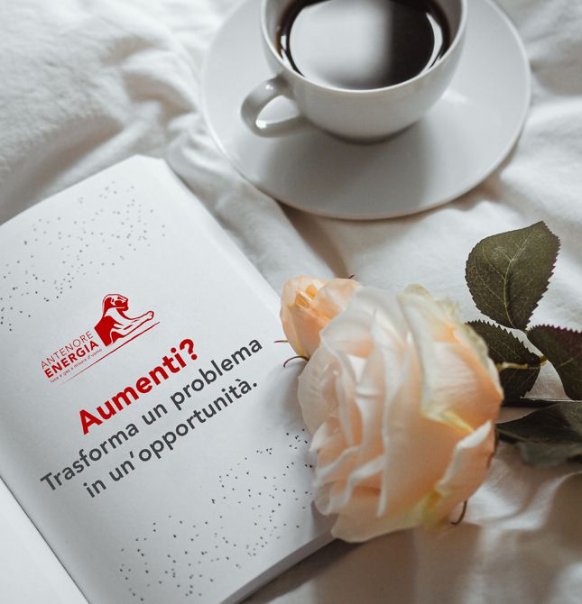 Tazza di caffè accanto a libro con scritta "Aumenti? Trasforma un problema in un’opportunità" e rosa bianca su lenzuolo