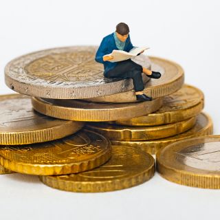 Miniatura uomo seduto su monete impilate legge giornale, concetto aumento bollette e gestione rincari energetici