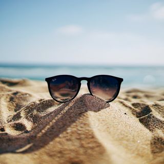 Occhiali da sole neri su sabbia dorata in spiaggia, sfondo mare e cielo estivo, concetto di caldo e risparmio energetico