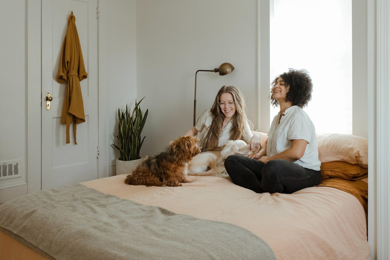 Due donne sorridenti su letto matrimoniale con cane e gatto, ambiente domestico luminoso, atmosfera rilassata e conviviale