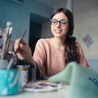 Donna sorridente con occhiali lavora a un progetto creativo in studio, circondata da pennelli e colori, ambiente domestico...