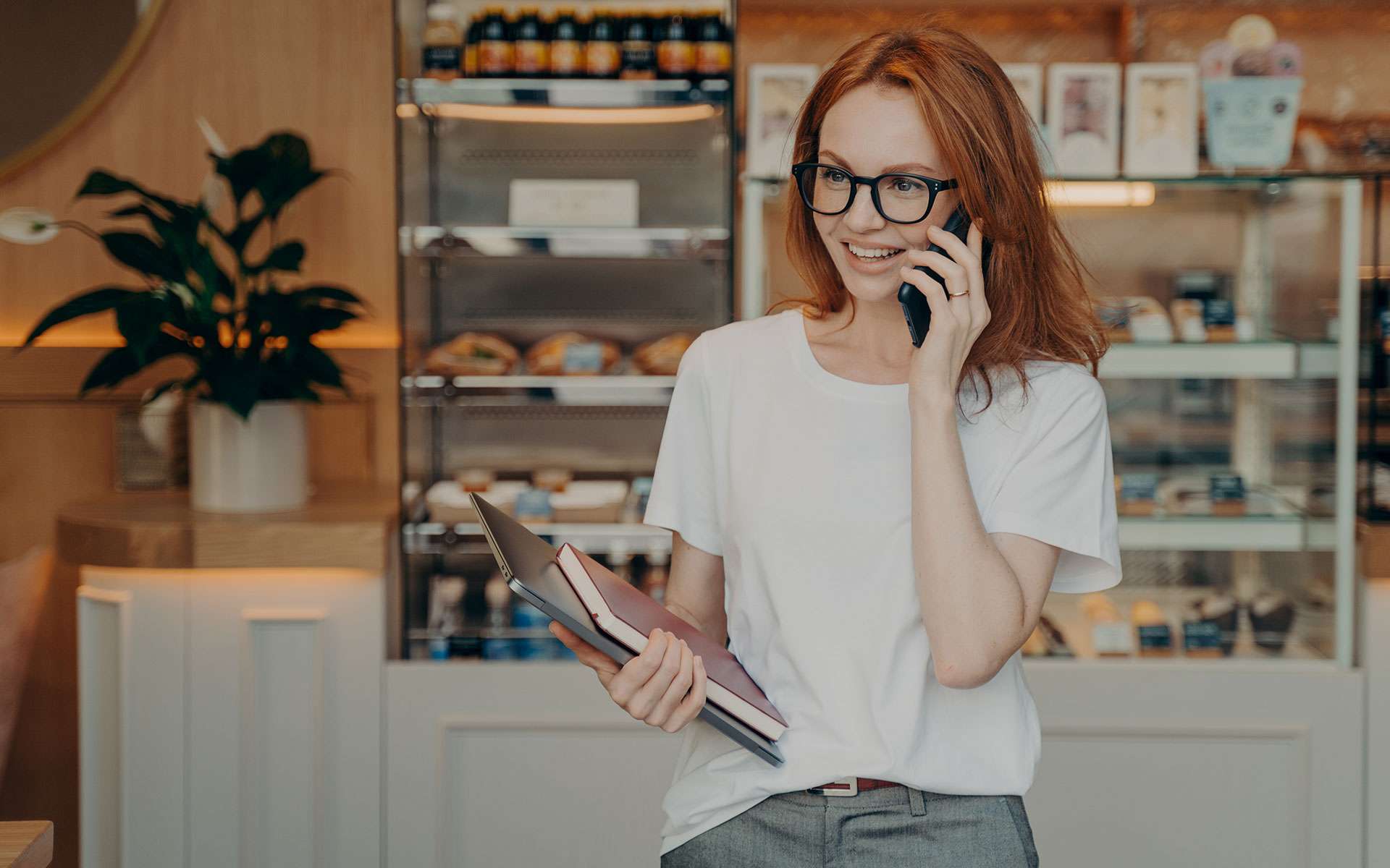 Donna sorridente al telefono in caffetteria moderna, tiene quaderni e indossa occhiali, atmosfera accogliente e professionale