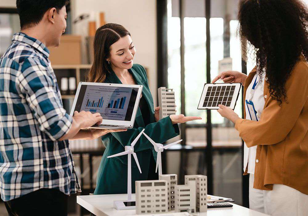 Tre persone discutono di energia rinnovabile con grafici, modellini di edifici, pale eoliche e pannello solare in ufficio