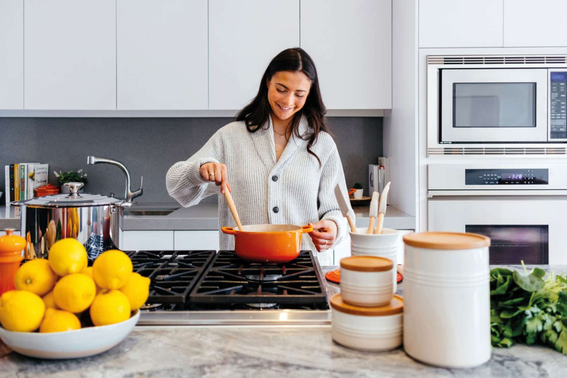 Donna cucina in cucina moderna con pentola arancione su fornello a gas, limoni freschi e contenitori in ceramica sul piano